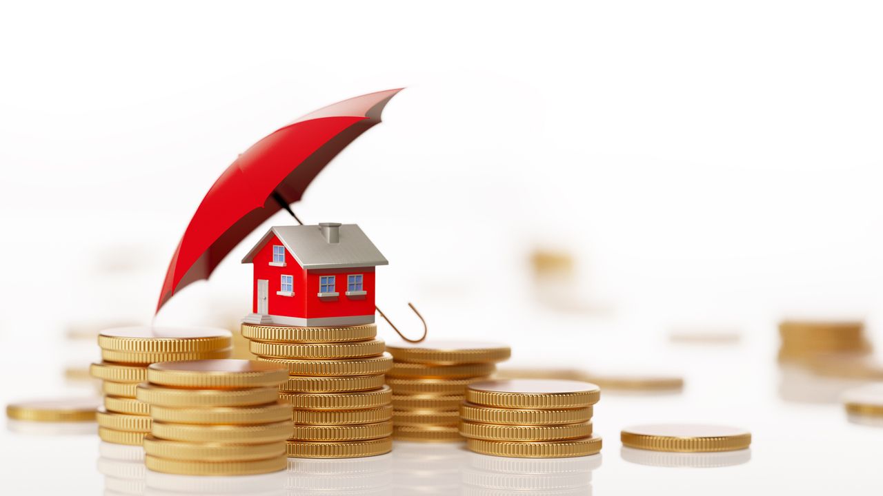 Red toy house is sitting behind a coin stack under a red umbrella. Horizontal composition with selective focus and copy space. Real Estate and Insurance Concept.